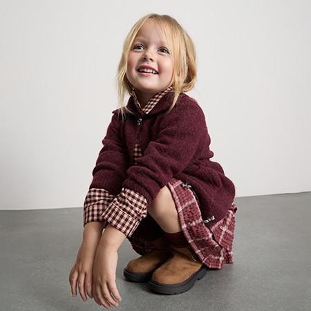 Young child wearing burgundy coat with checked collar over matching dress and brown boots.