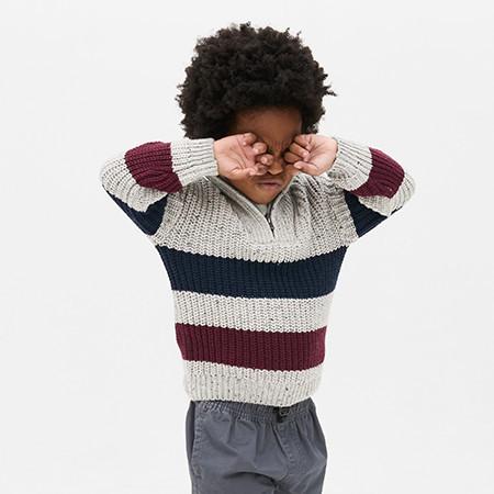Child in striped knitted jumper with burgundy, navy and cream bands rubbing eyes with both hands.