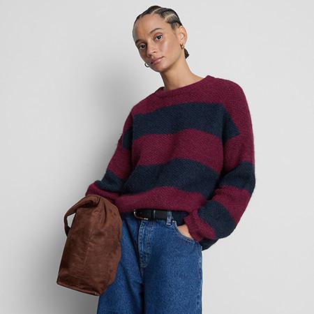 Person wearing burgundy and navy striped knit jumper with blue jeans, holding a brown bag.