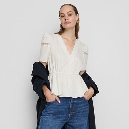 Woman wearing cream V-neck peplum blouse with lace detail and blue jeans, black jacket draped over arms.