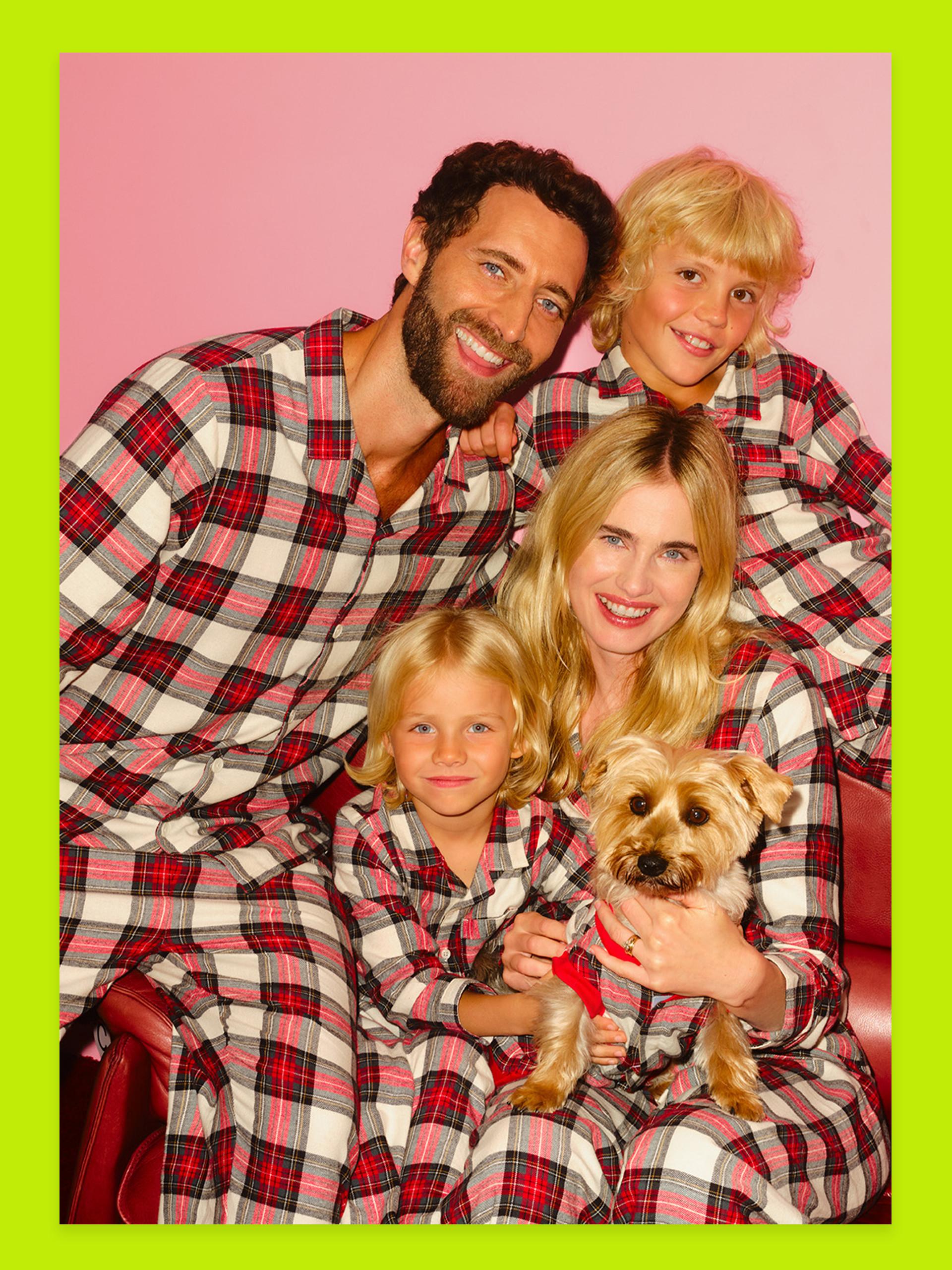 Family wearing matching red and white plaid pyjamas with small dog against pink background.