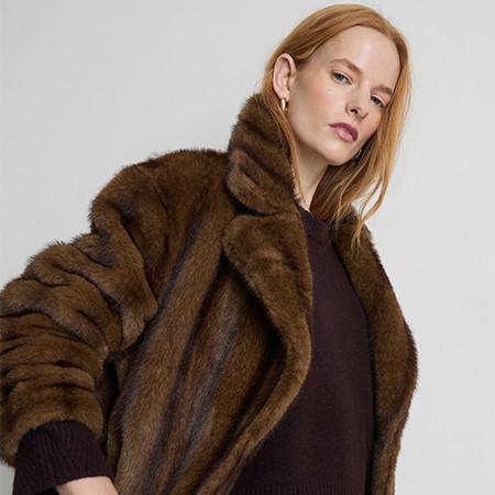 Person wearing luxurious brown faux fur coat over dark purple sweater against grey background.