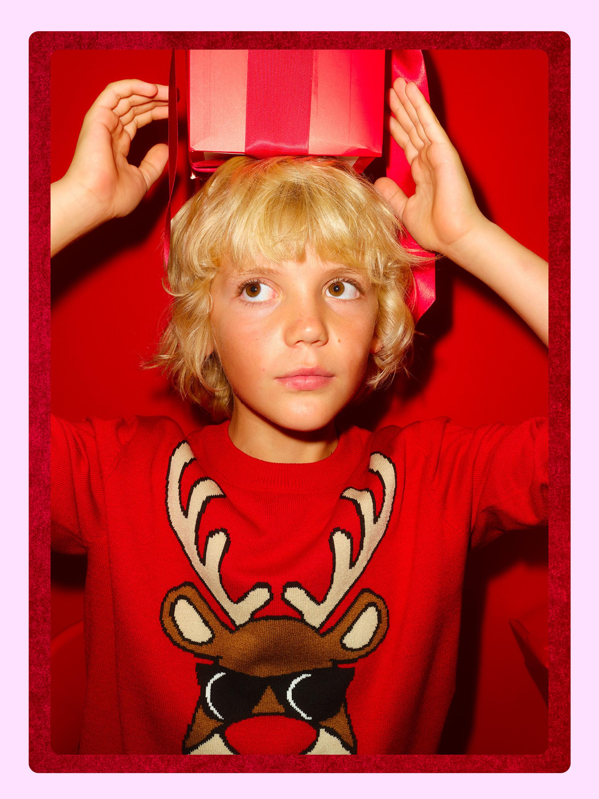 Child wearing red Christmas jumper with reindeer design balancing a striped gift box on head.