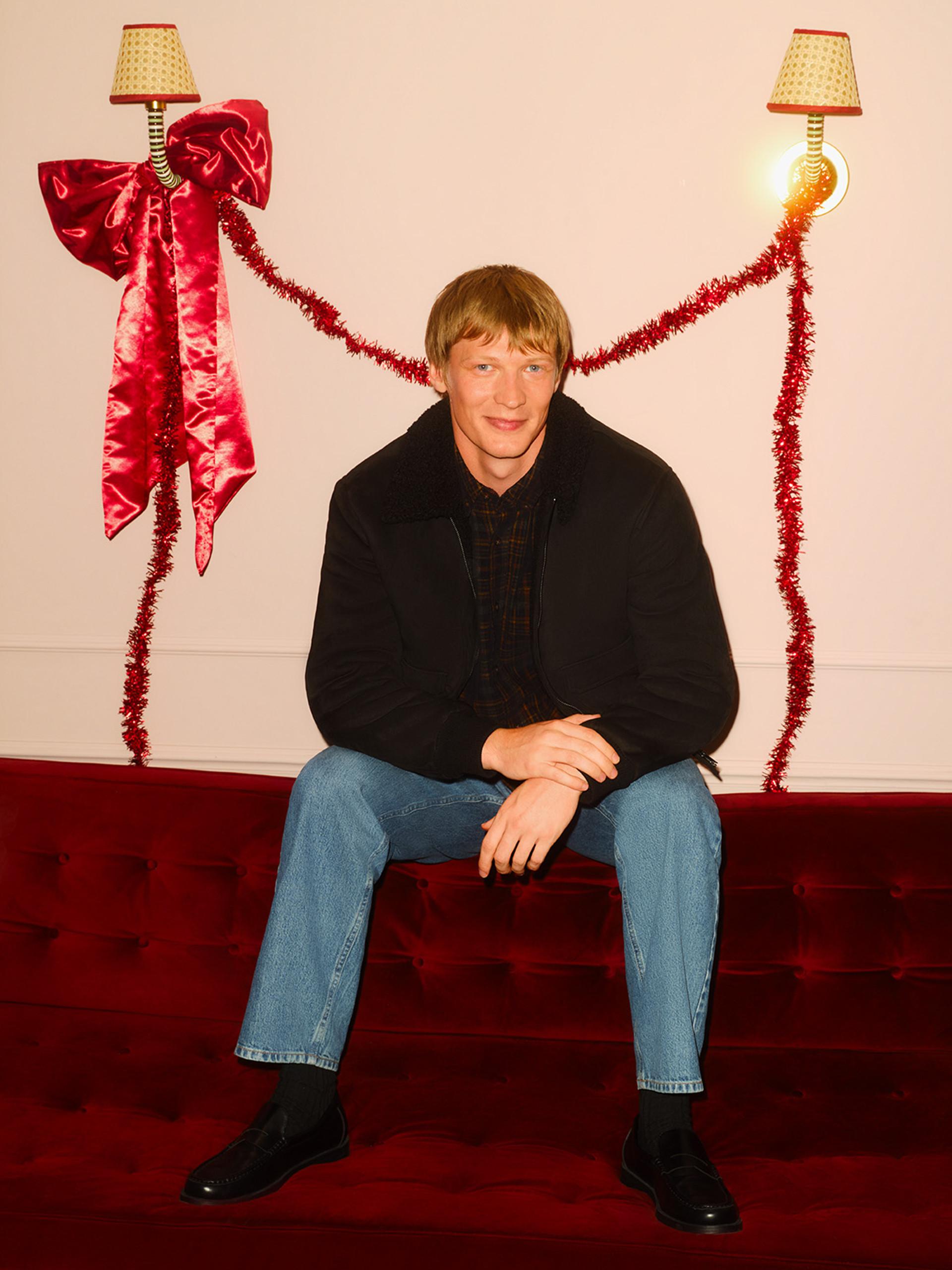 Person sitting on red velvet sofa wearing black jacket and jeans with festive red tinsel decorations and lamps behind.