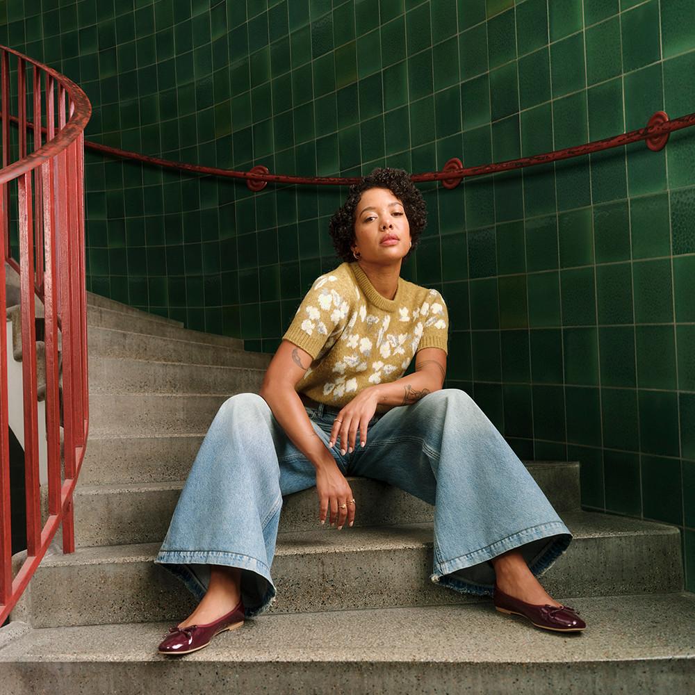 Person wearing floral yellow knitted jumper, wide-leg jeans and burgundy flats sitting on concrete stairs with green tiled walls.