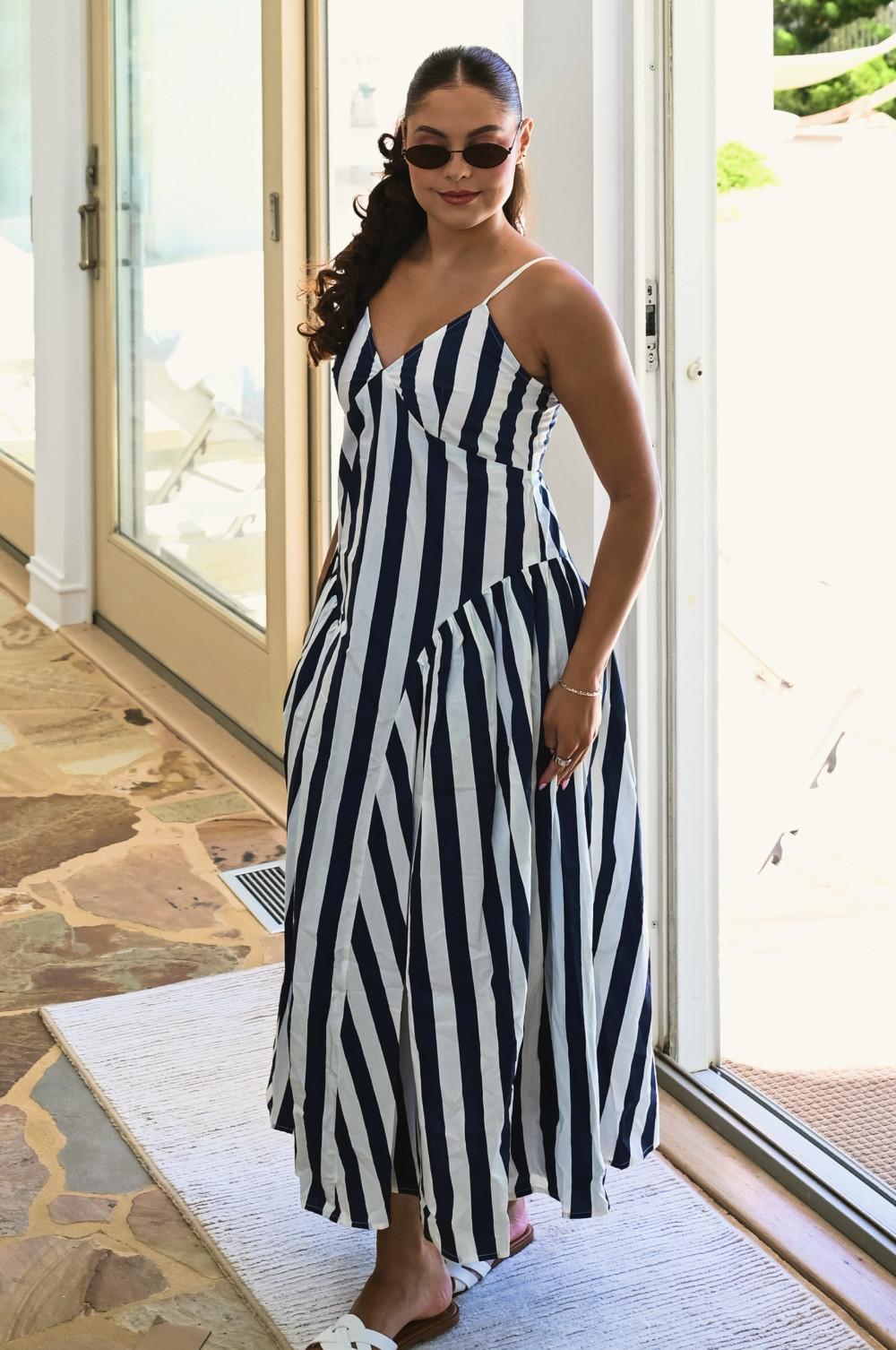 Model wearing navy and white striped maxi dress with sunglasses, standing in a doorway.