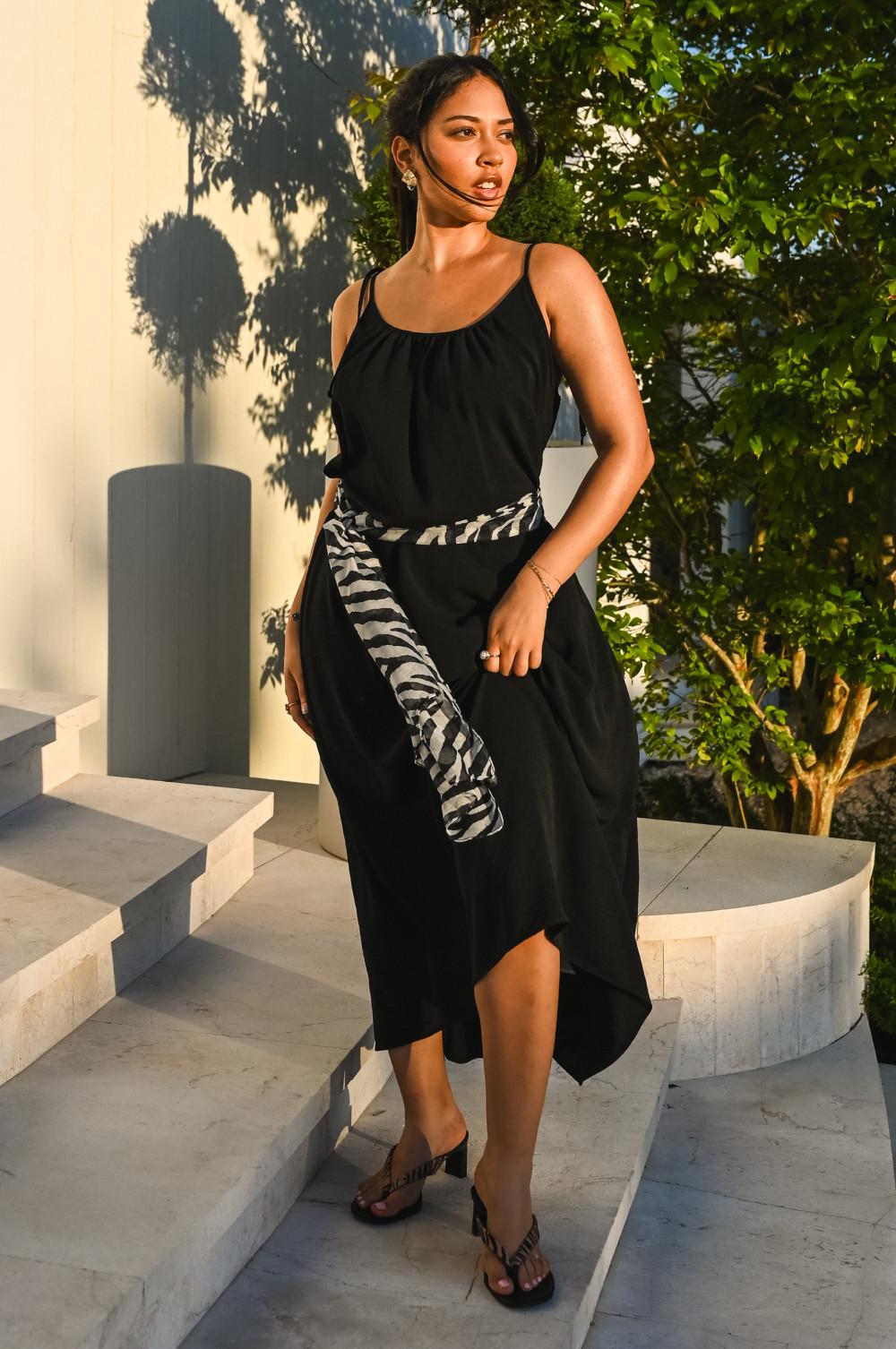 Model wearing black sleeveless midi dress with zebra print jumper tied at waist, standing outdoors.