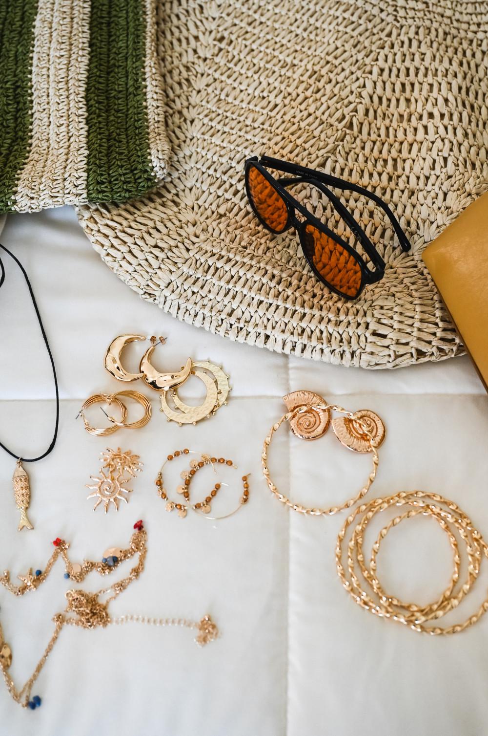 Flat lay of gold jewellery including earrings, necklaces and bracelets with sunglasses on woven mat.