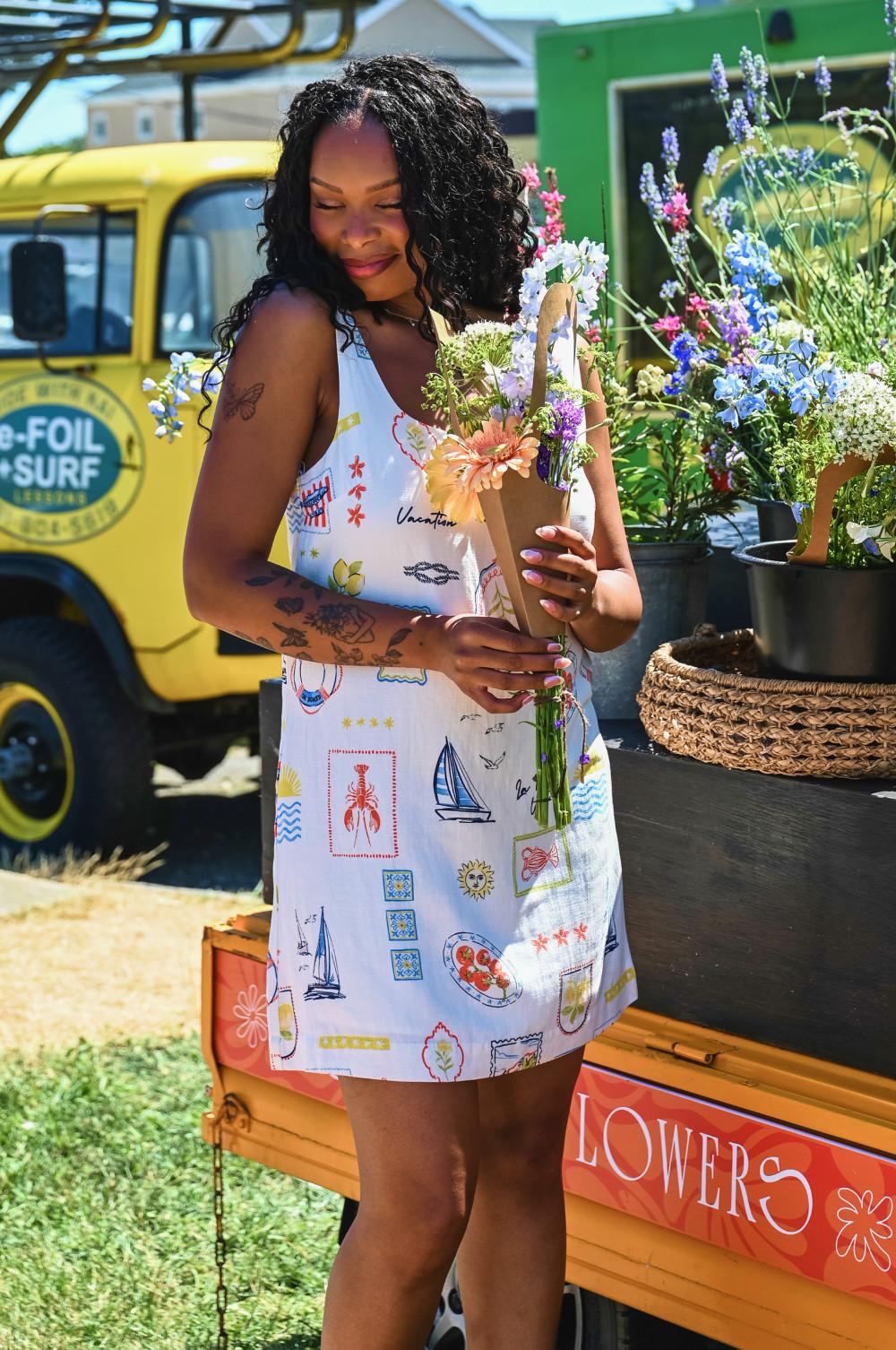 Model wearing white sleeveless dress with colourful holiday motif print, holding bouquet of wildflowers.