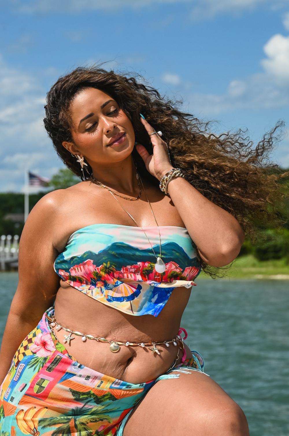 Model wearing a colourful tropical print bikini with gold jewellery, posing outdoors by water.
