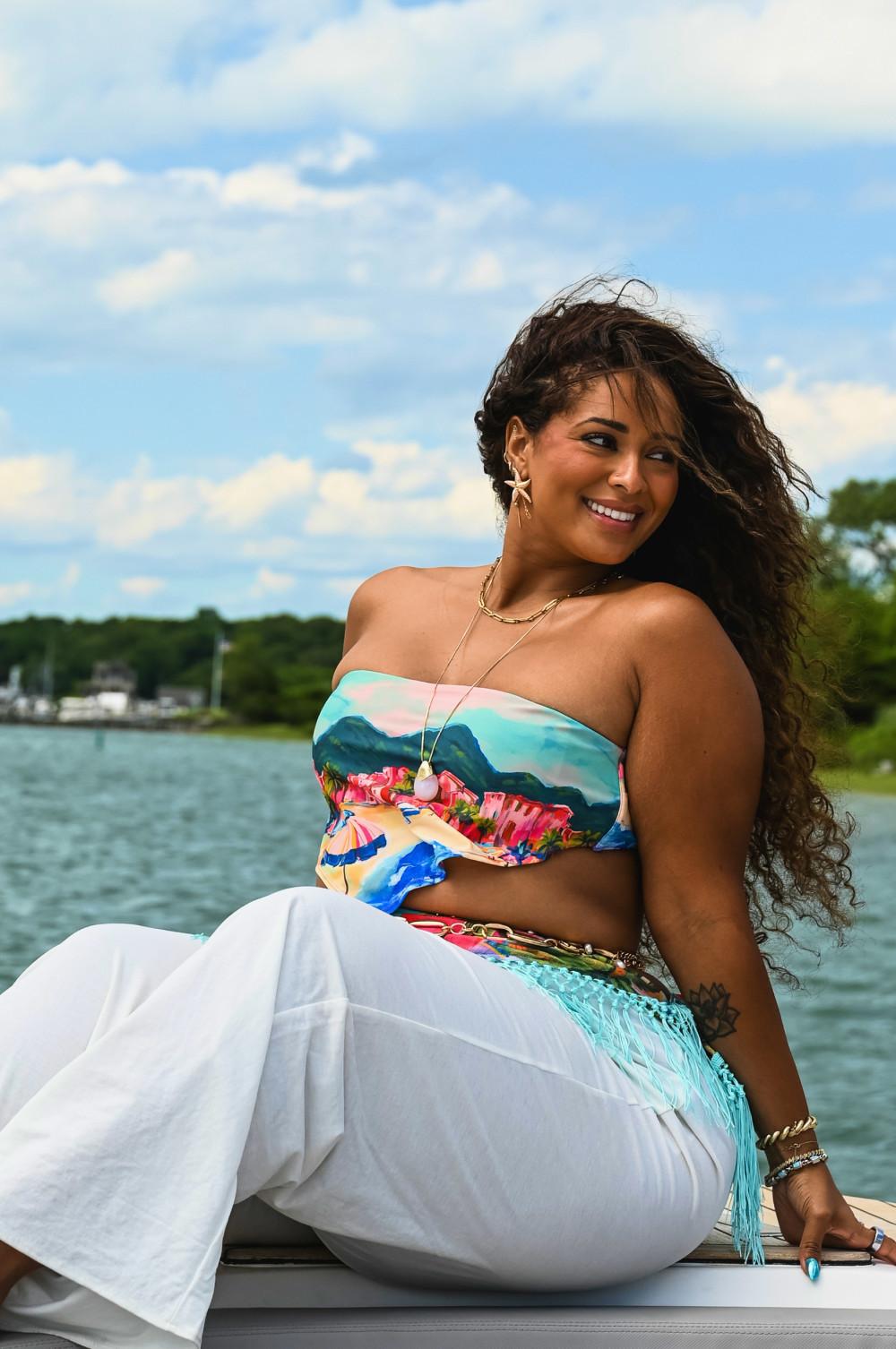 Model wearing colourful bandeau bikini top and white trousers sitting by waterside on sunny day.