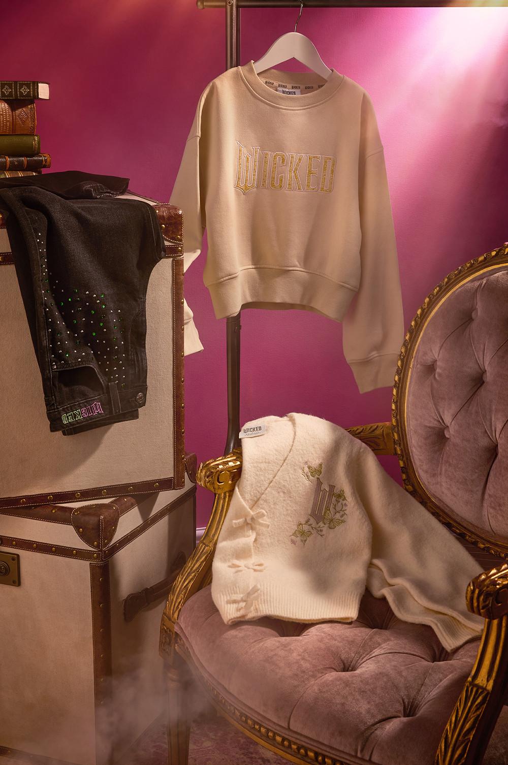 Beige Wicked sweatshirt hanging on display with coordinating knitwear on a velvet chair against a pink backdrop.