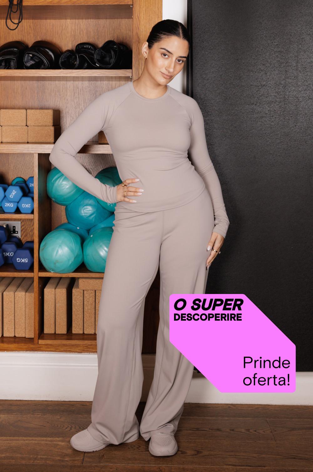 Persoană purtând îmbrăcăxminte sport bej într-un studio de fitness cu echipamente sport vizibile pe rafturile din lemn.