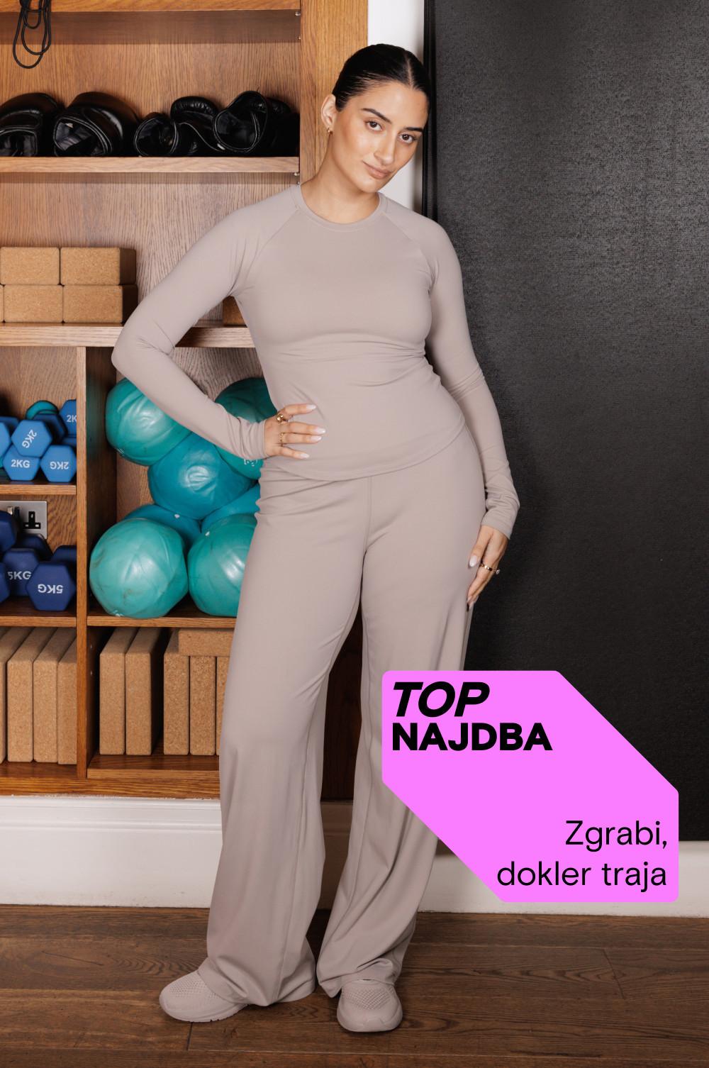 Oseba v bež športnih oblačilih v fitnes studiu z vadbeno opremo, vidno na lesenih policah.