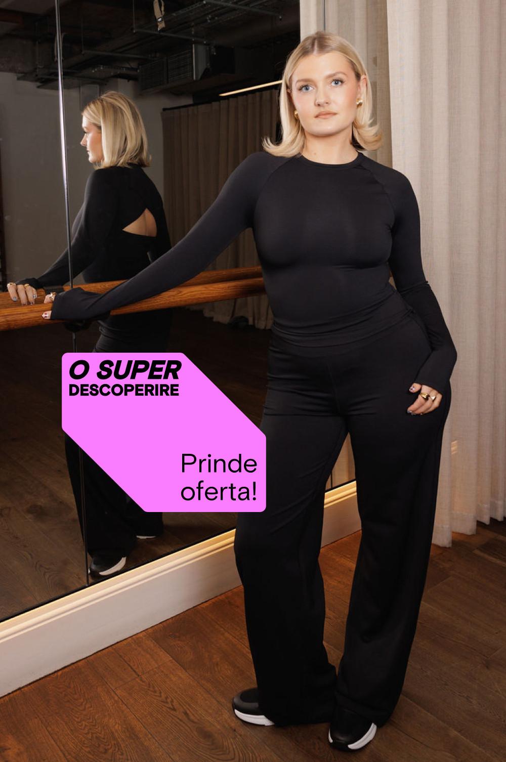 Persoană purtând un top negru cu mânecă lungă și pantaloni mulați, lângă o oglindă cu o etichetă promoțională roz „SUPER DESCOPERIRE”.