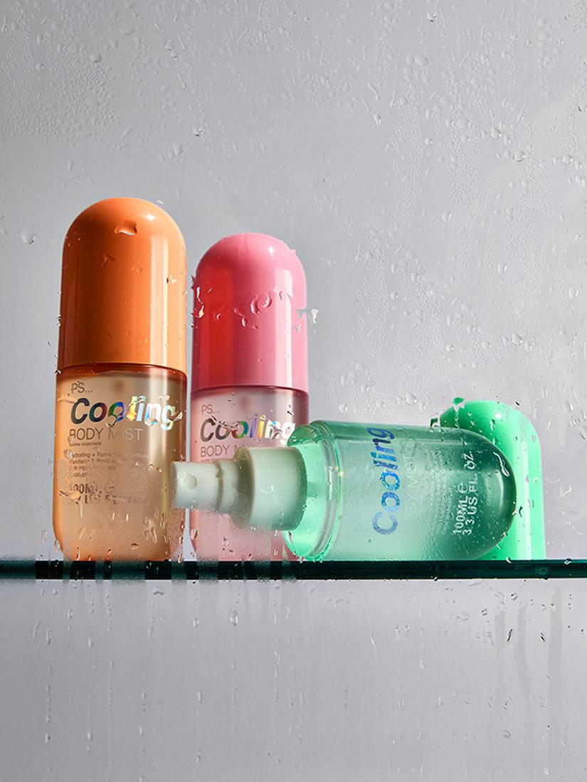 Three PS Cooling body mist bottles in orange, pink and green with water droplets on glass shelf.