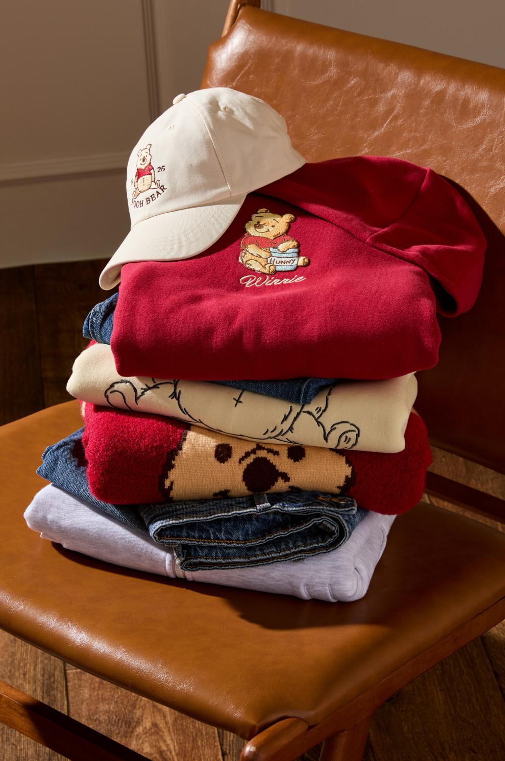 Stack of Winnie the Pooh themed clothing including a white cap, red hoodie, and various garments on a leather chair.