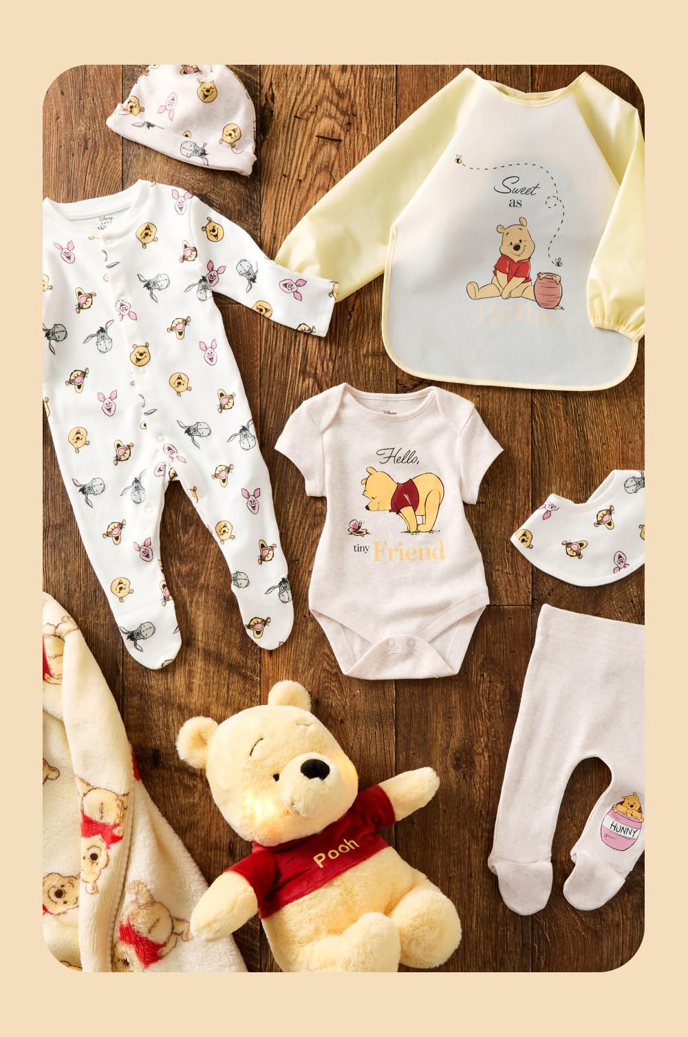 Winnie the Pooh baby clothing collection with bodysuit, sleepsuit, bib, hat and soft toy displayed on wooden surface.