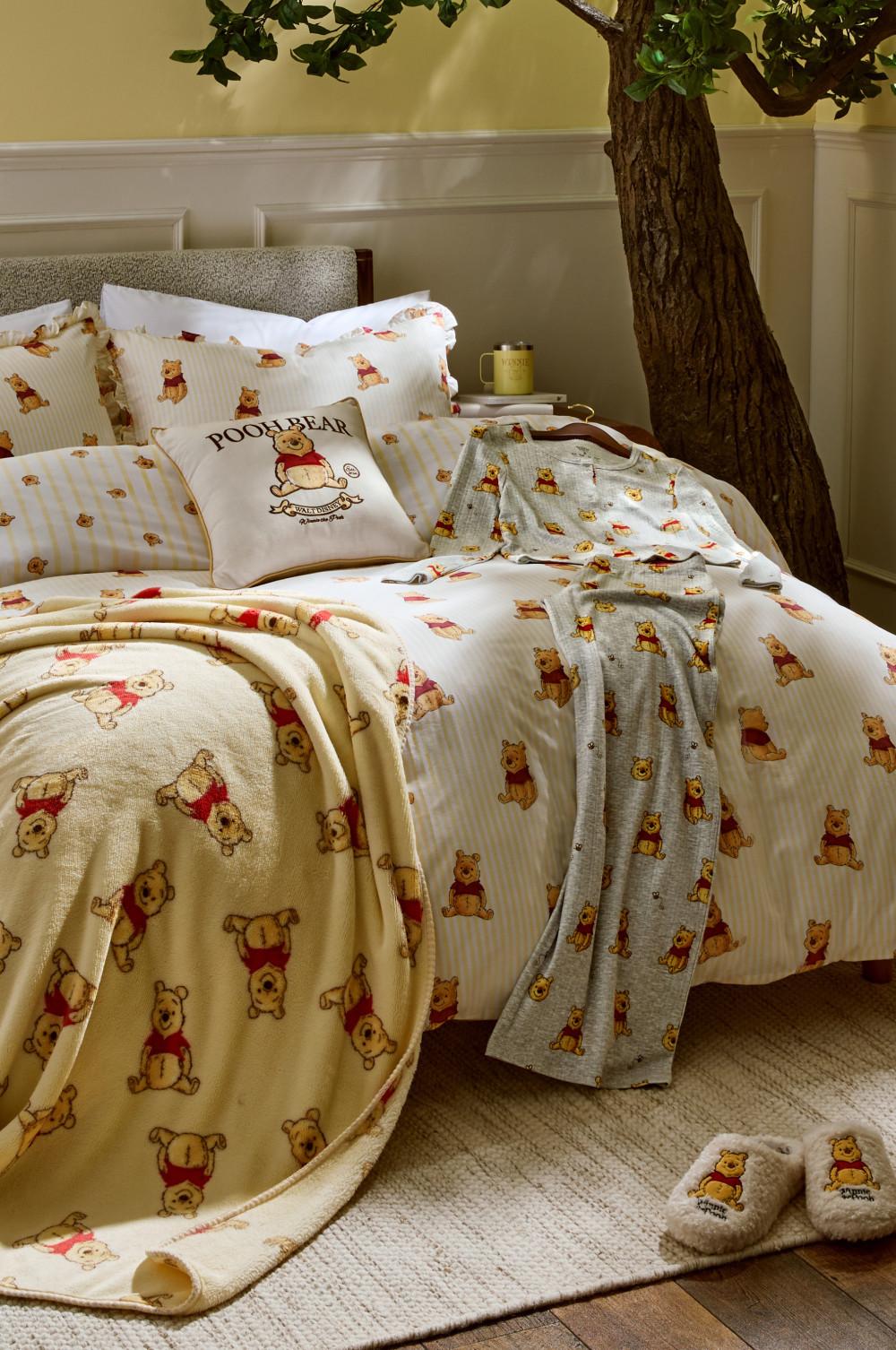 Winnie the Pooh themed bedding set with matching blanket, cushion, pyjamas and slippers beneath a decorative indoor tree.