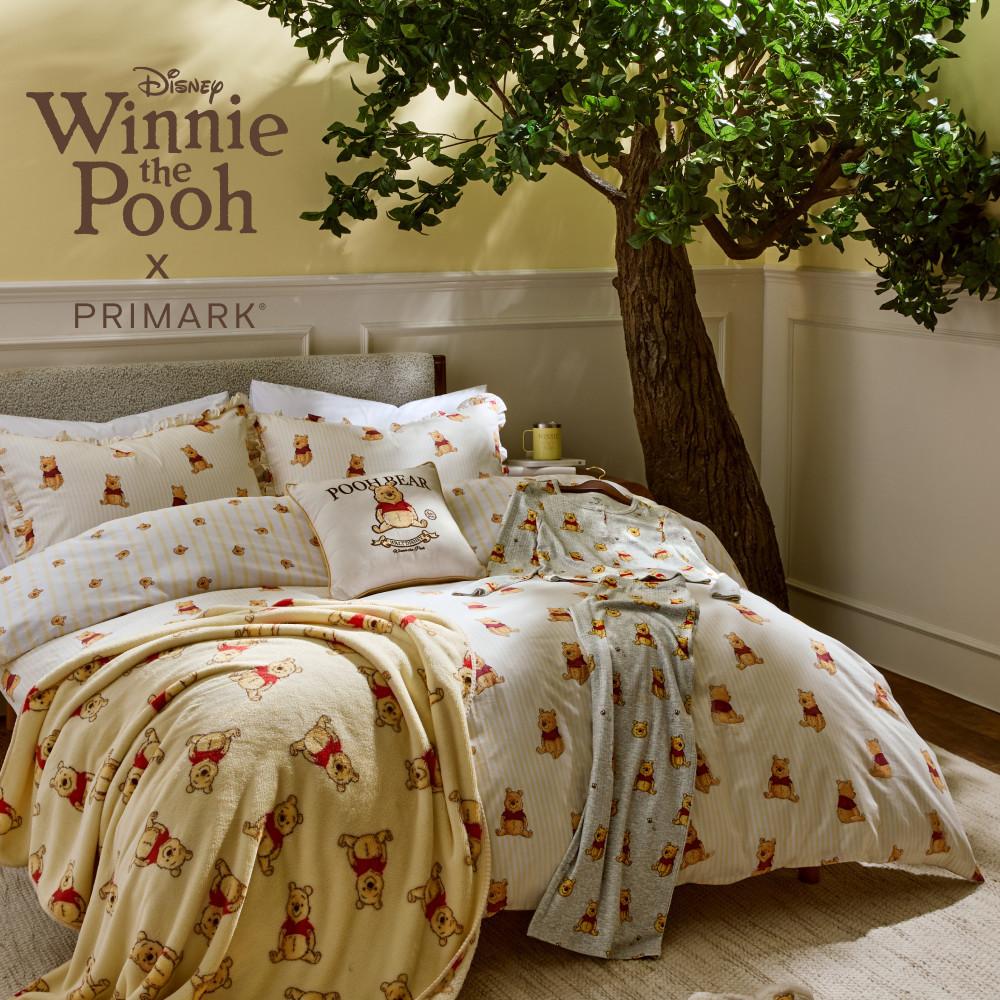 Ropa de cama de la colección Disney Winnie the Pooh x Primark con edredón, mantas y cojines estampados junto a un árbol decorativo.