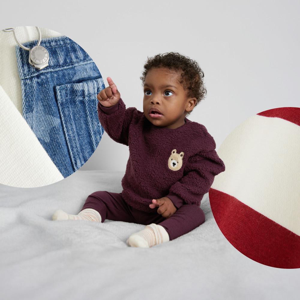 Enfant tendant la main vers un tissu en denim et portant un pull bordeaux à motif ourson avec un pantalon assorti et des chaussettes blanches.