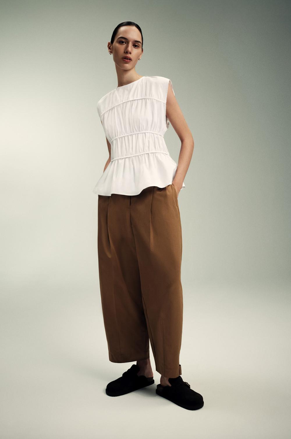 Top blanco péplum sin mangas, pantalón marrón de pernera ancha y mocasines negros en un entorno minimalista.