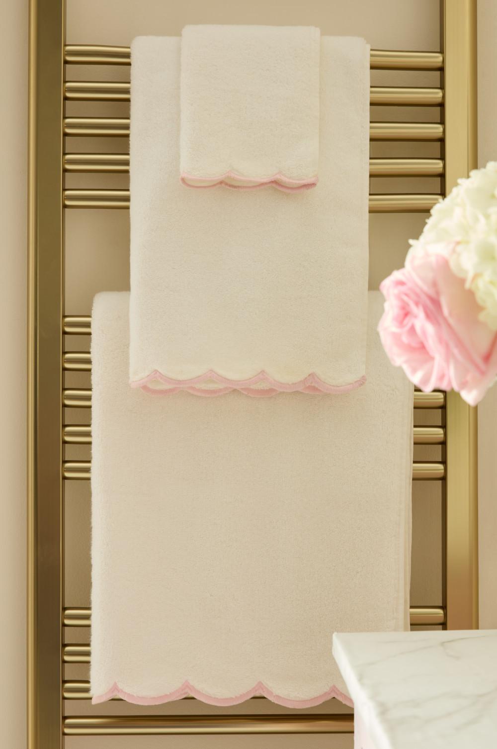Toallas blancas con ribete festoneado rosa colgadas en un toallero dorado calefactado, con flores decorativas a un lado.