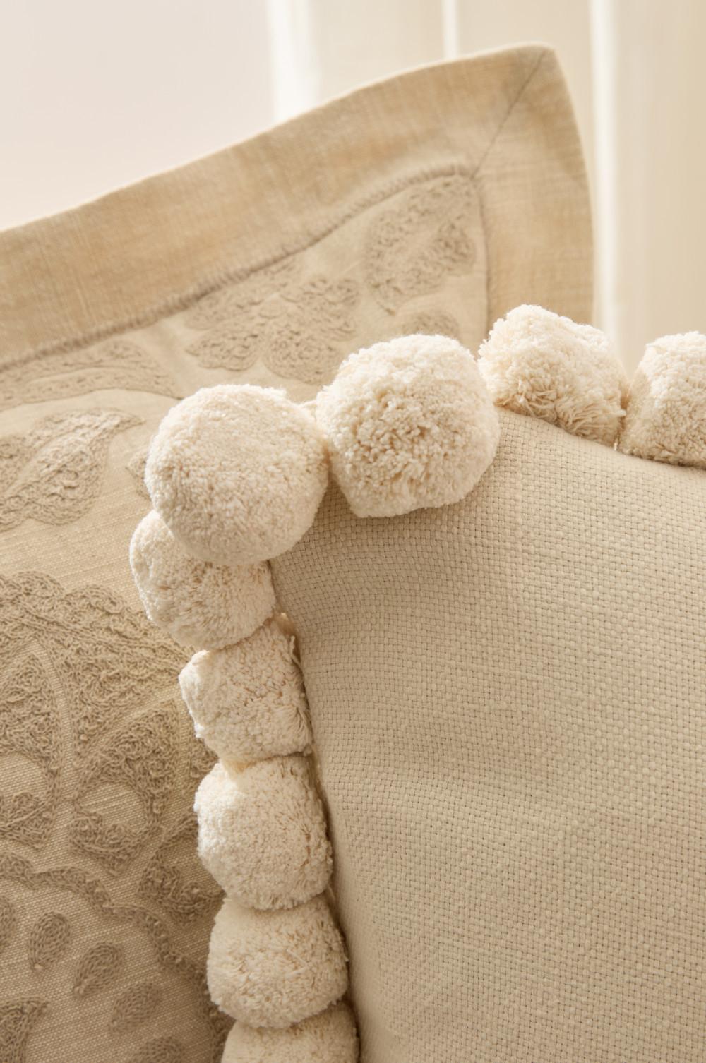 Cojín color crema con ribete de pompones junto a otro bordado en tonos neutros.