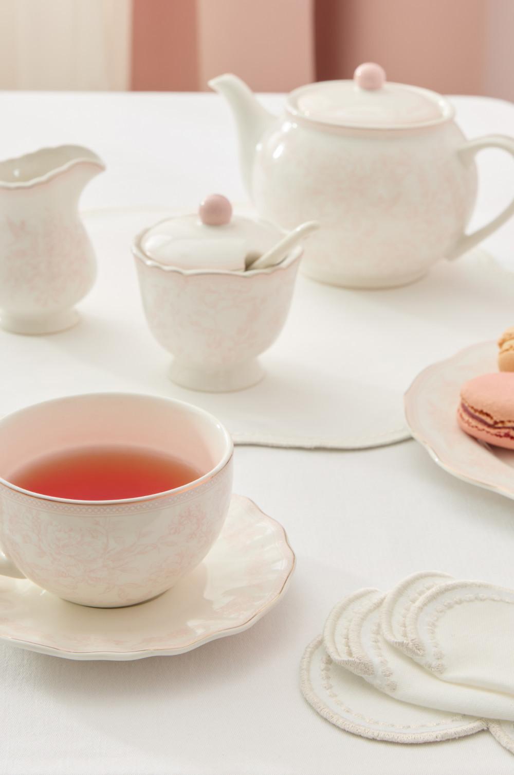 Elegante juego de té en blanco y rosa con tetera, azucarero, taza de té y pastas de té sobre un mantel blanco.