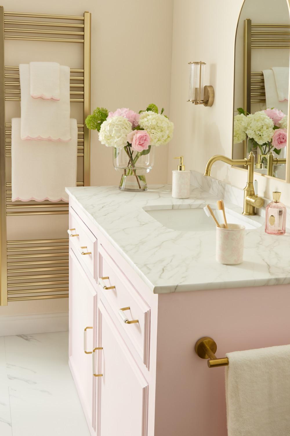 Baño elegante con tocador rosa, encimera de mármol, grifería de latón y flores frescas en un jarrón.