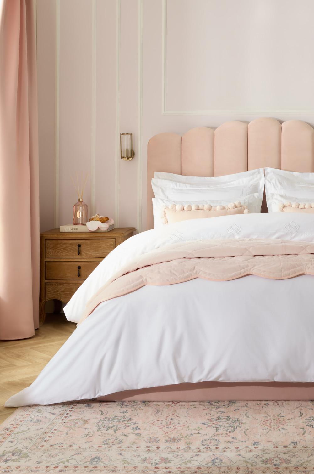 Dormitorio elegante con cabecero tapizado en rosa, ropa de cama blanca, mesita de noche de madera y detalles en rosa empolvado.