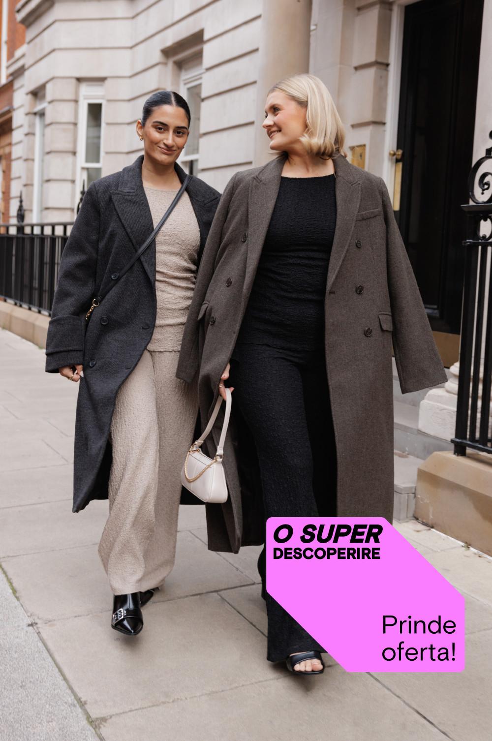 Două femei care se plimbă împreună purtând paltoane de iarnă elegante și ținute elegante, în fața unei clădiri. Etichetă promoțională roz cu textul „SUPER DESCOPERIRE”.
