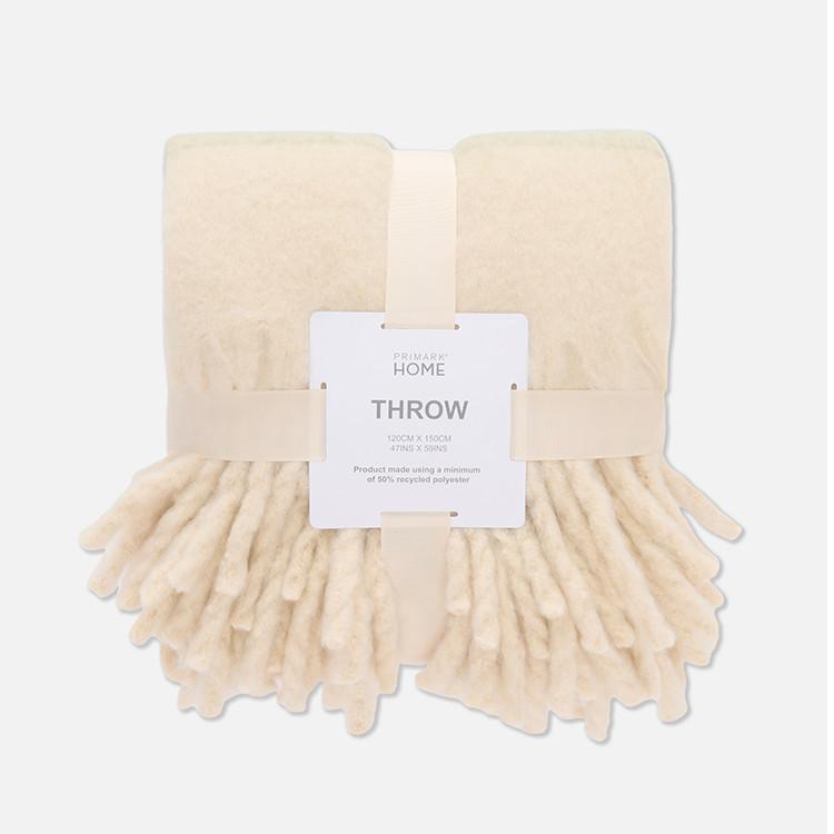 Cream coloured Primark Home throw blanket with tasselled edge, folded with product label attached.