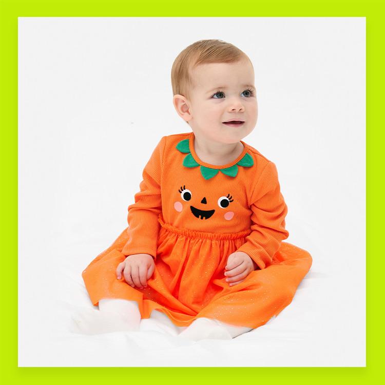 Baby wearing bright orange pumpkin costume dress with smiling face design and green collar detail.