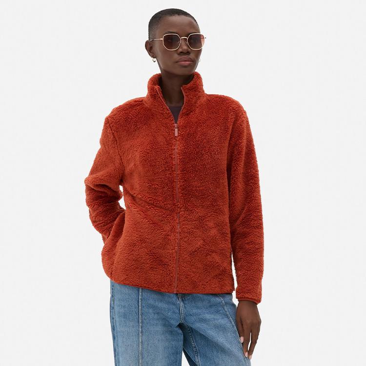 Person wearing rust-coloured fluffy fleece jacket with high collar, blue jeans and round sunglasses.