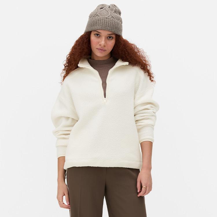 Person wearing cream fleece half-zip pullover with grey beanie and brown trousers.