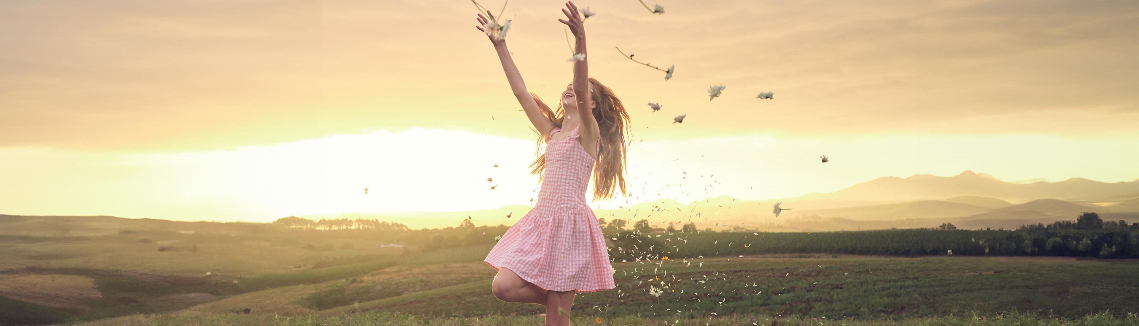 A girl in a pink dress joyfully throws leaves in the air in a scenic field at sunset.