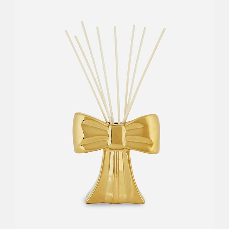 Gold bow-shaped reed diffuser with thin white reeds extending from the top of the decorative container.