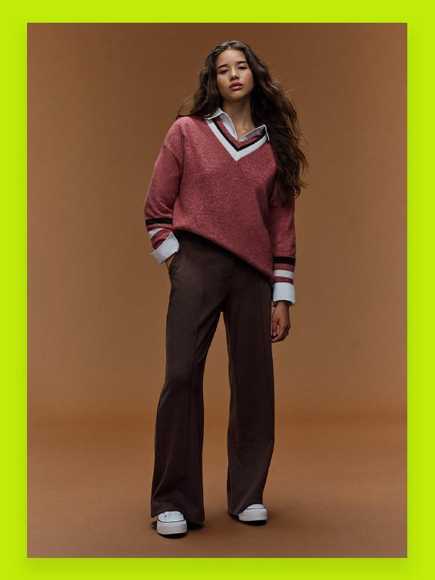 Person wearing pink v-neck jumper with striped collar over brown wide-leg trousers and white trainers.