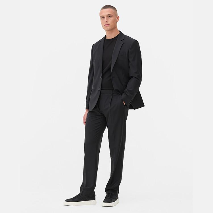 Person wearing black formal suit with matching t-shirt and trainers, standing with hand in pocket.
