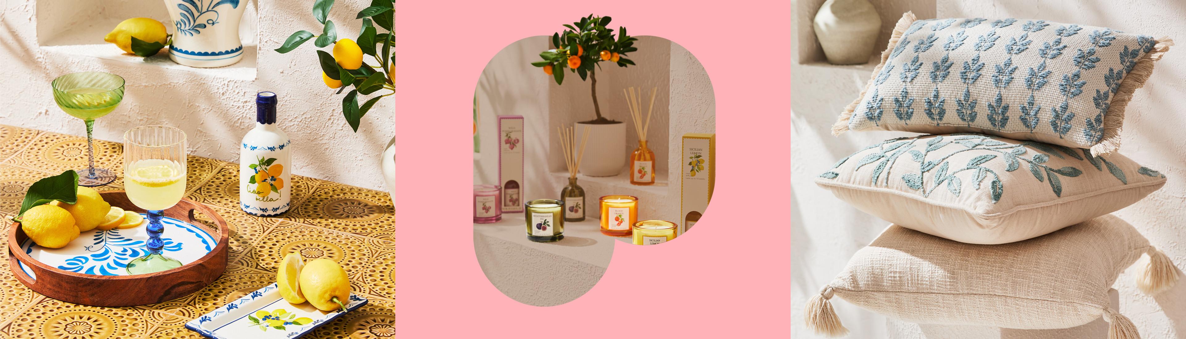 Tischdekoration im Zitronen-Look mit Flasche, Gläsern, frischen Zitronen und dekorativem Teller auf einer gewebten Matte. Ein Stapel dekorativer Kissen mit blauer Blumenstickerei und Quasten in Naturtönen.