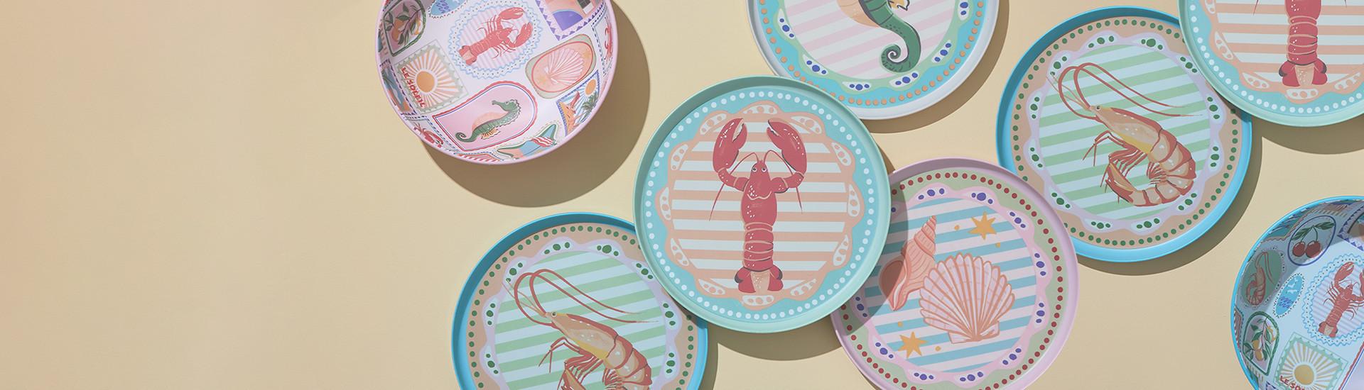 Decorative coastal-themed plates with sea creature designs including lobster, shrimp, seahorse and shell motifs on pastel striped backgrounds.