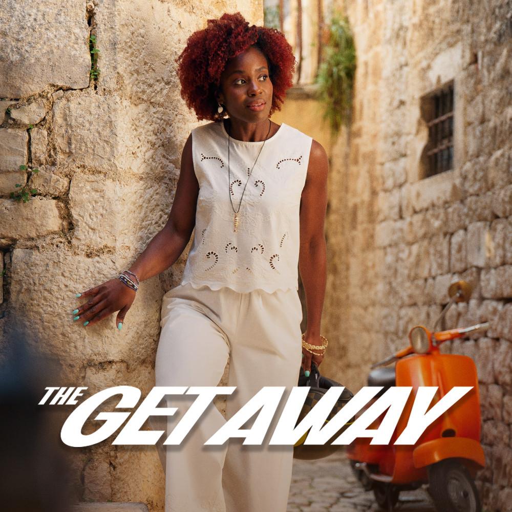 Woman leaning against stone wall wearing white sleeveless broderie anglaise top and trousers with jewellery.