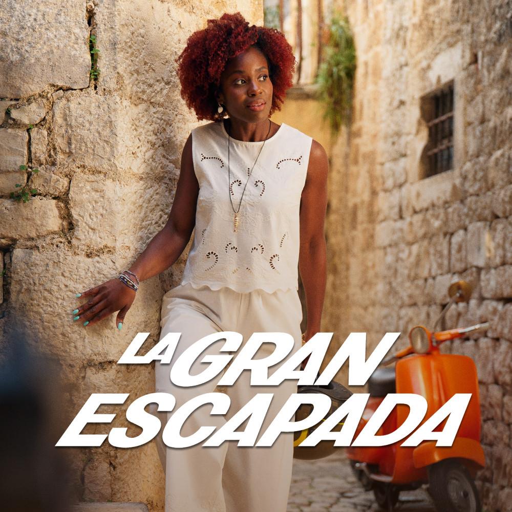 Woman leaning against stone wall wearing white broderie anglaise sleeveless top and cream trousers.