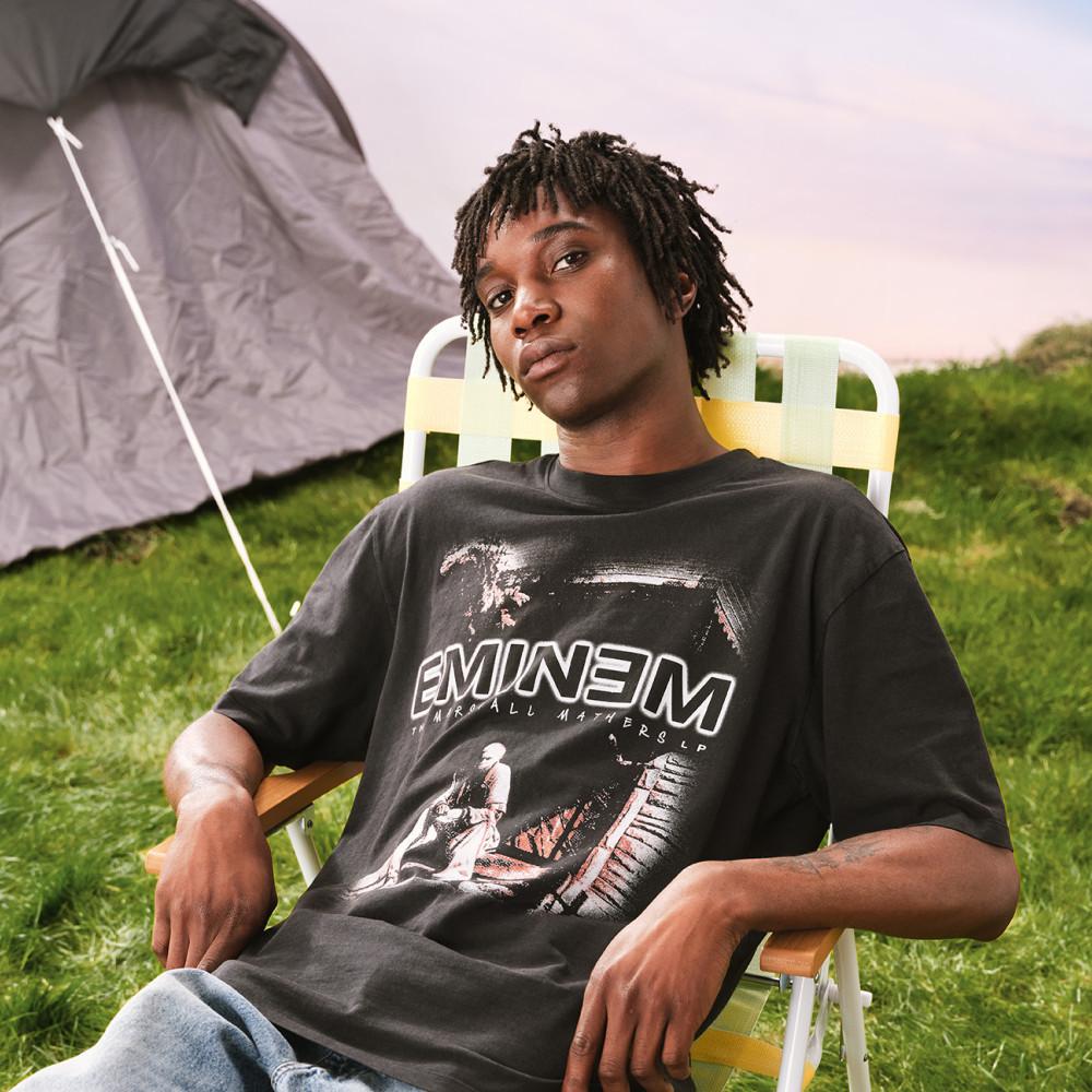 Person relaxing on camping chair wearing black Eminem graphic t-shirt outdoors near tent.