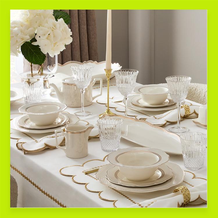 Elegant table setting with white and gold dinnerware, crystal glasses and a tall candle, featuring white hydrangeas in a vase.