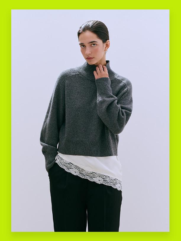 Grey knitted jumper with high neck over white lace-trimmed top, paired with black trousers.