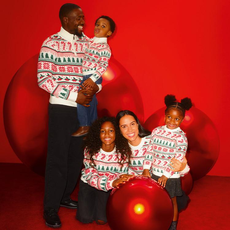 Family wearing matching Christmas jumpers with festive patterns against a bright red background.