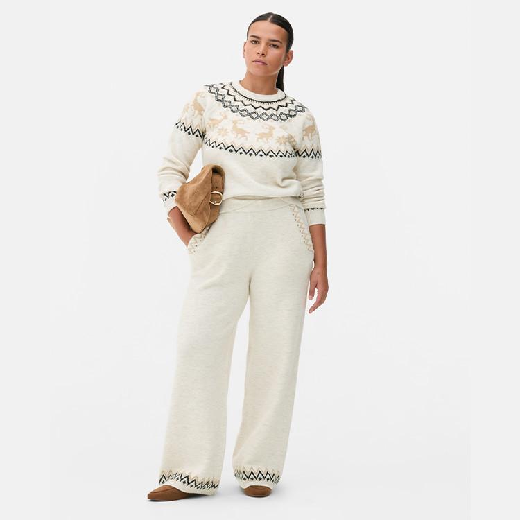 Person wearing cream Nordic pattern jumper and matching wide-leg trousers, holding a brown suede clutch bag.