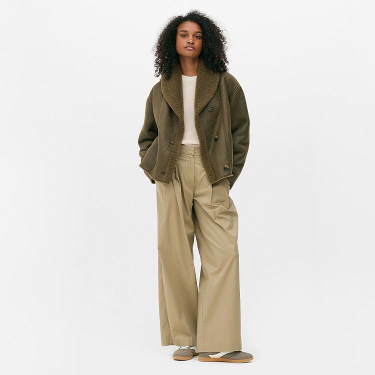 Person wearing olive green shearling jacket over white top with beige wide-leg trousers.